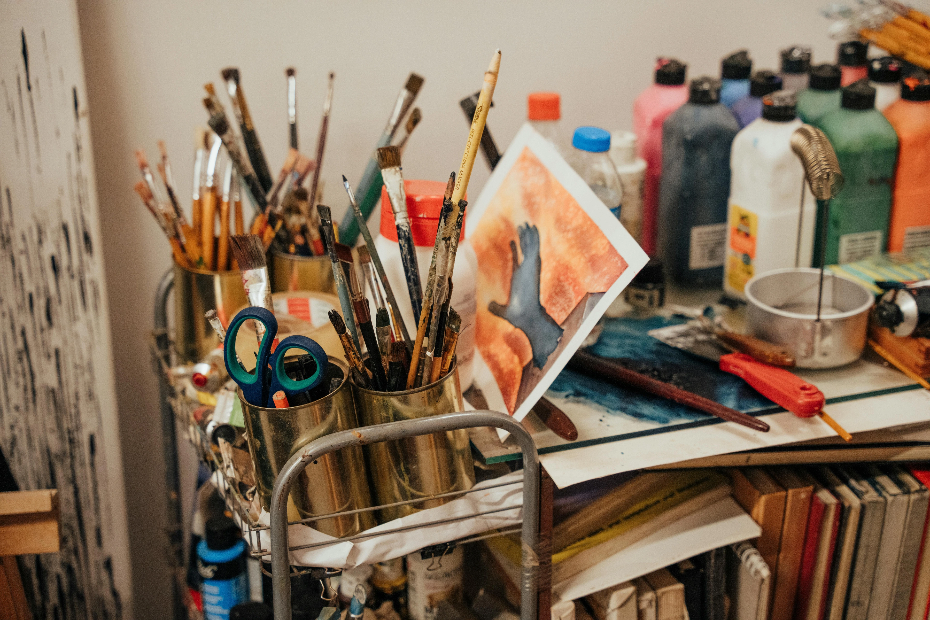 Close-up of a rolling cart filled with paintbrushes in metal containers, bottles of paint, and a small painting.