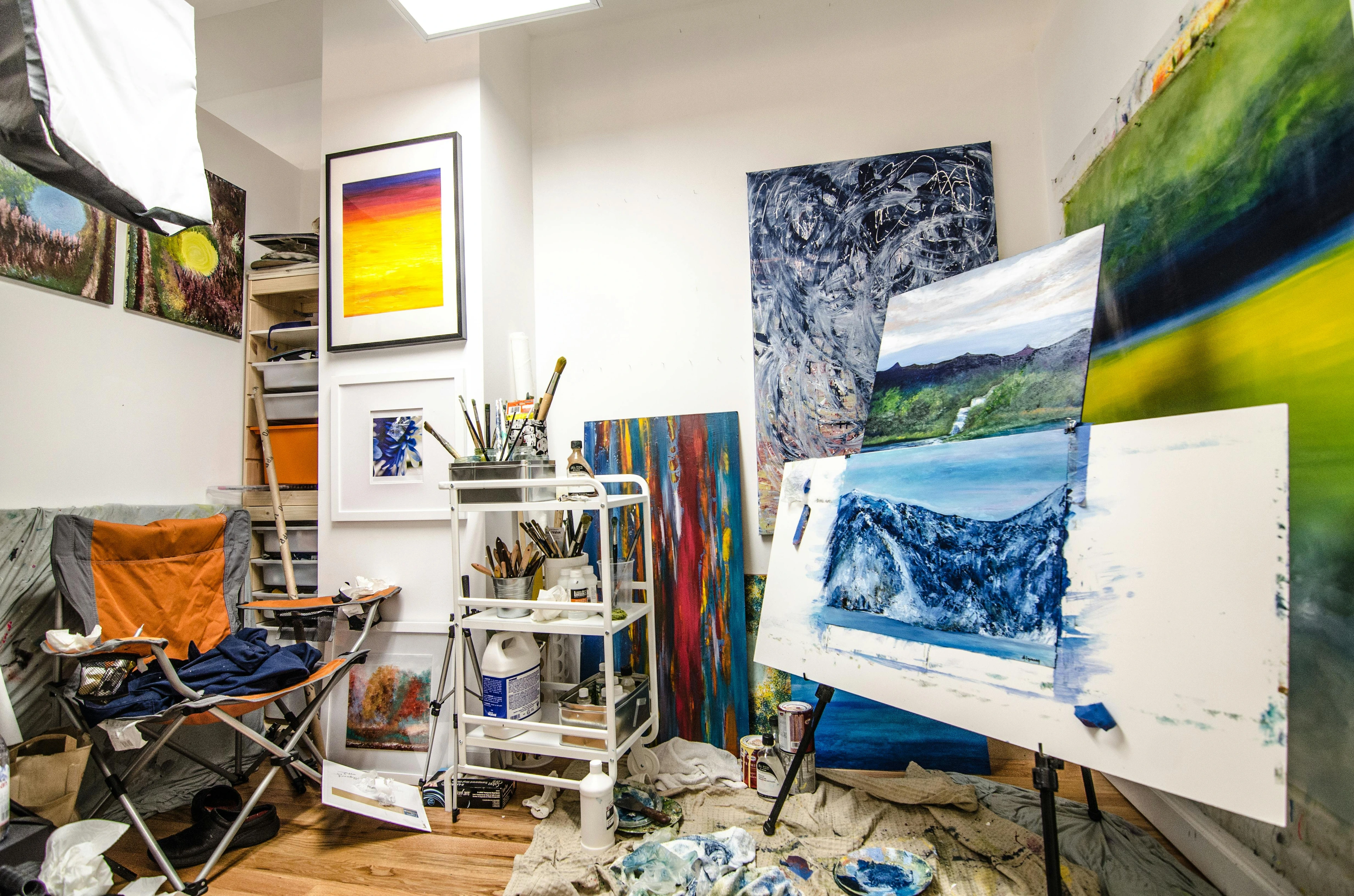 Photo of a cluttered art studio with paintings on easels and walls, art supplies, and furniture.