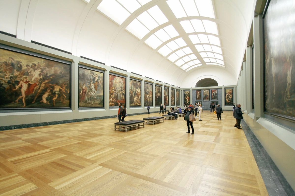 Long gallery in the Louvre Museum with numerous large framed paintings on the walls and visitors observing the art.