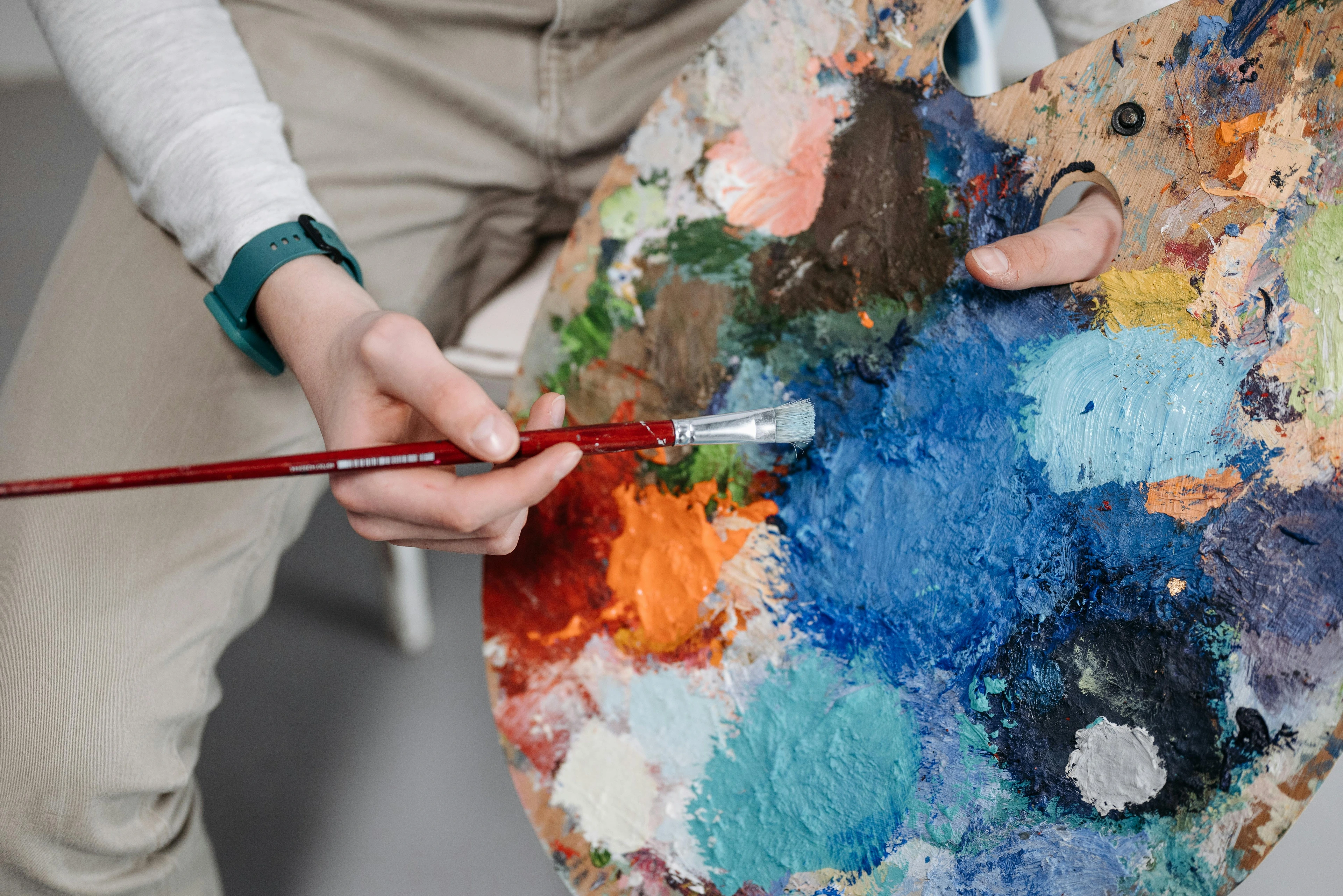 Artist's hands holding a paint palette with various colors and a paintbrush mixing blue paint.