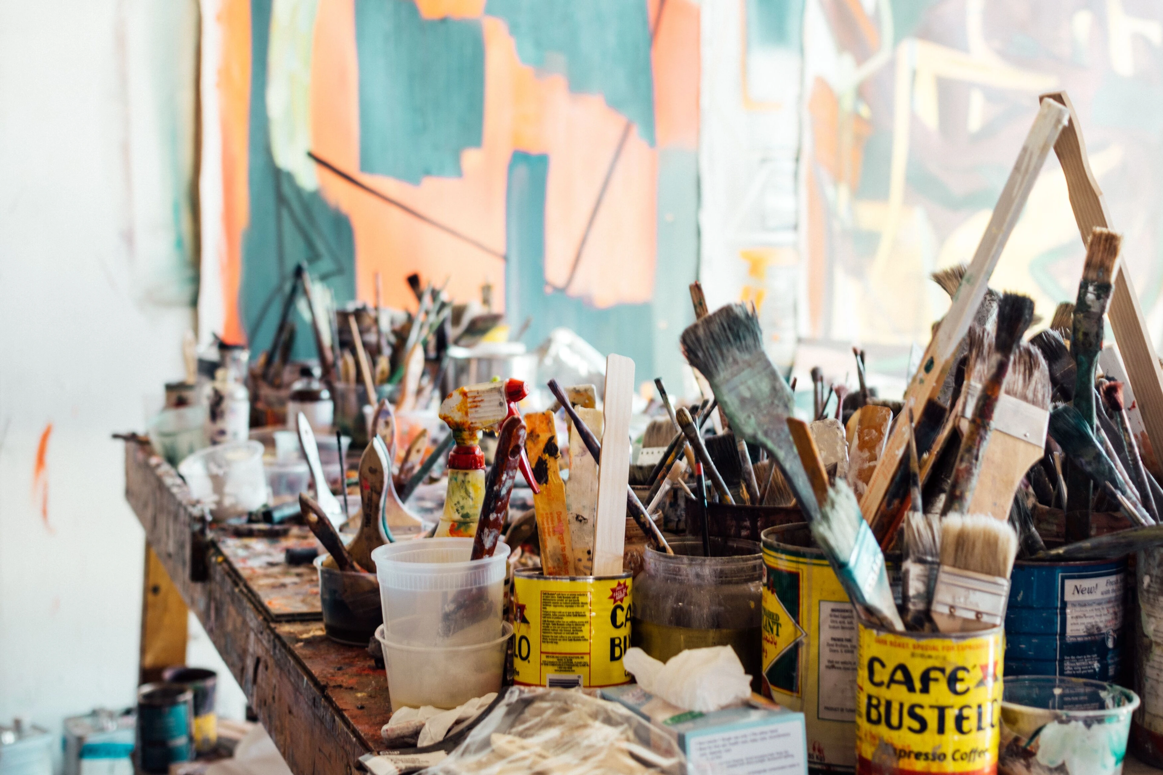 Cluttered artist's workbench with brushes, paints, and tools. Abstract painting visible in background.