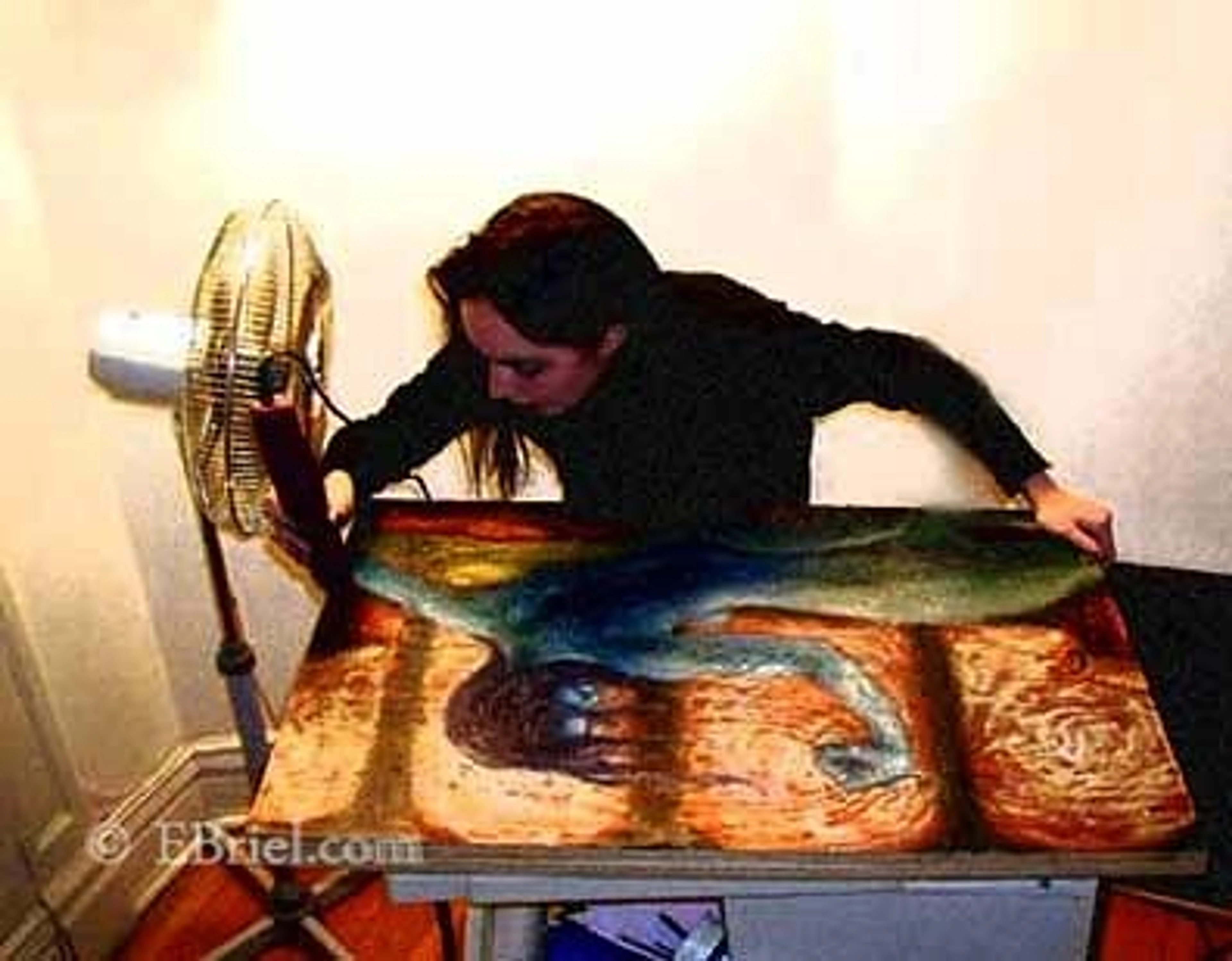 Artist working on an encaustic painting with a fan blowing on it.