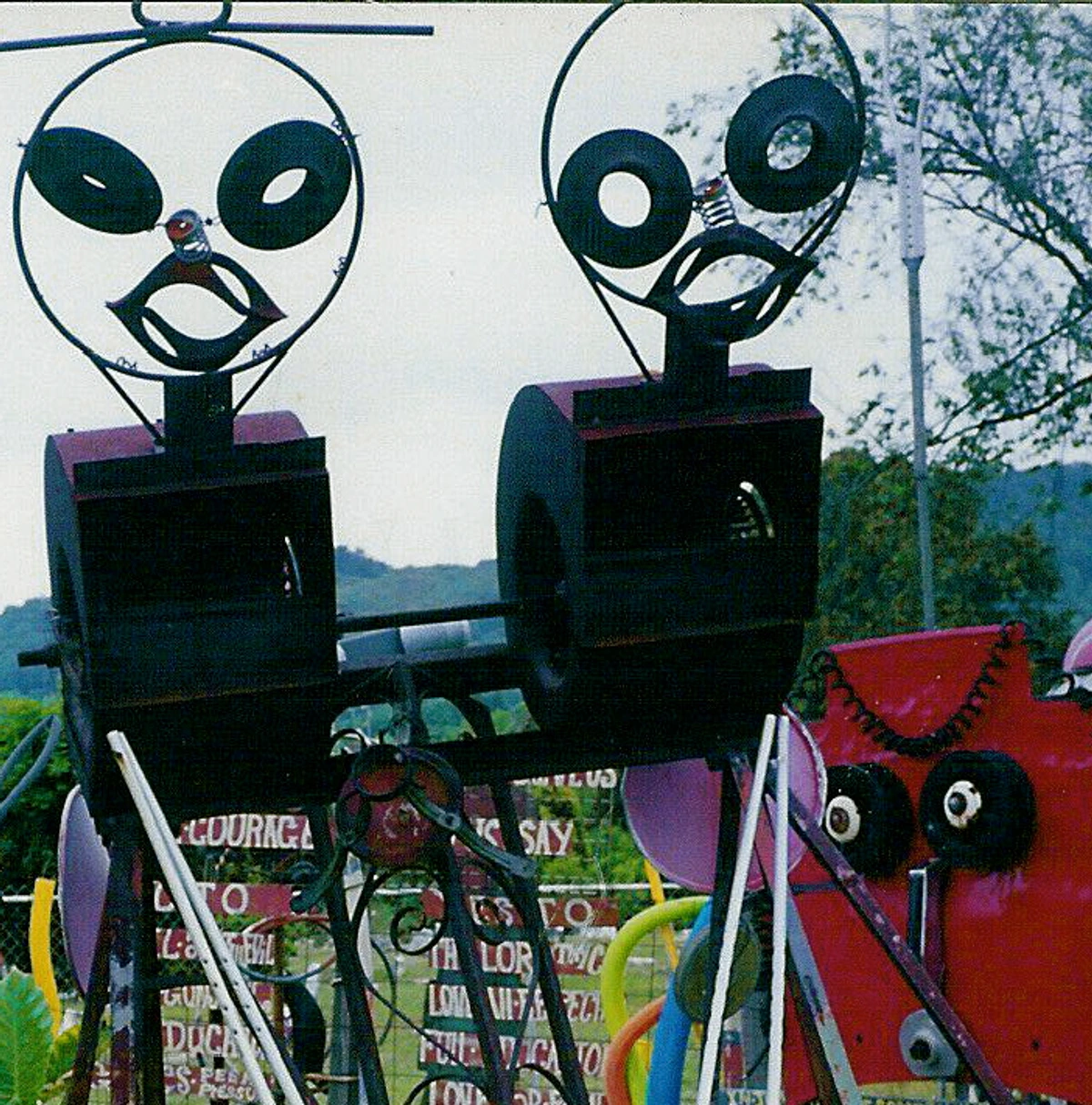 Two whimsical metal sculptures resembling aliens or robots, part of Joe Minter's African Village in America, with signs in the background.