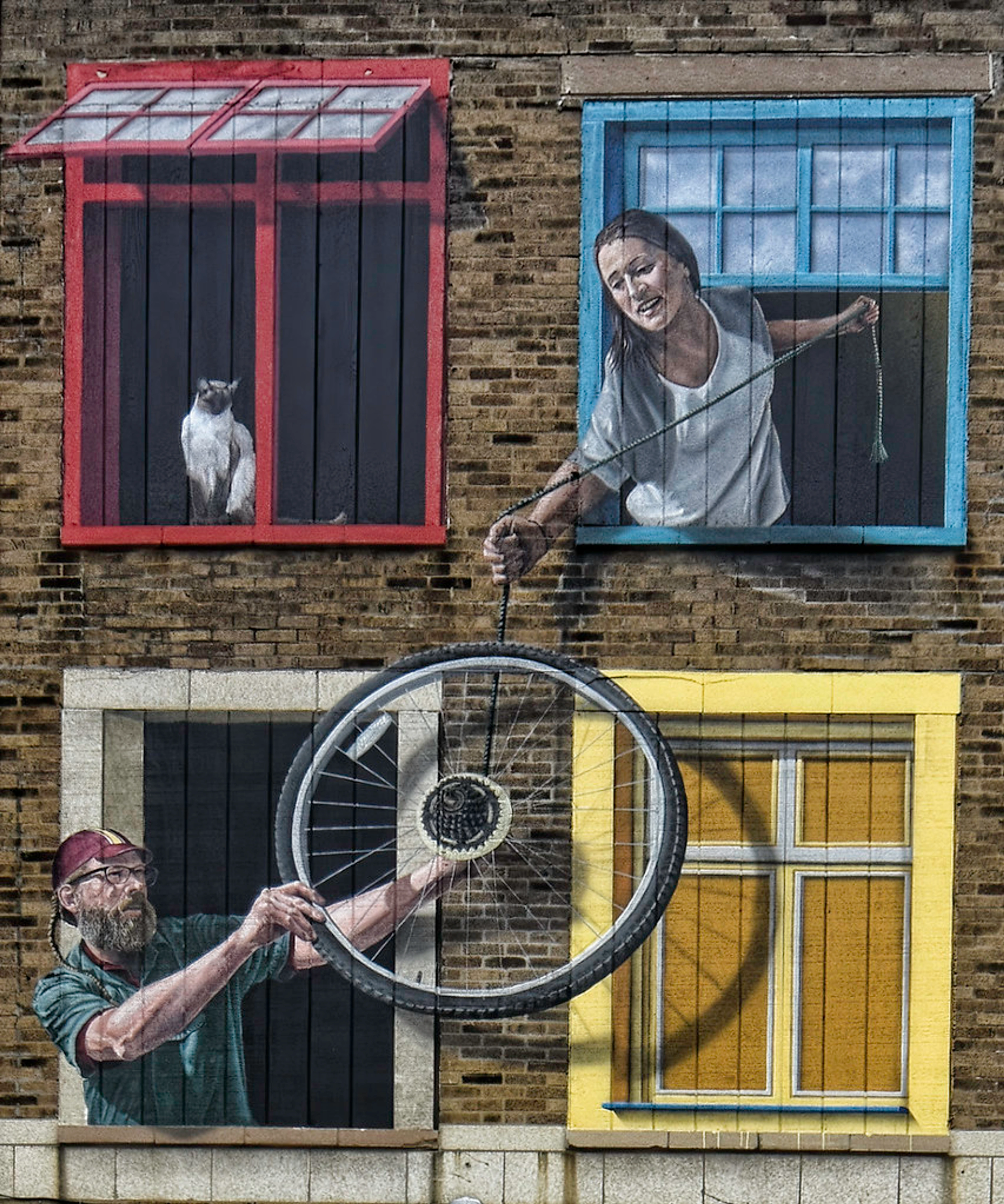 Trompe l'oeil mural on a brick building featuring a cat in a red window, a woman pulling a rope from a blue window, and a man holding a bicycle wheel from a white window.